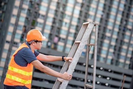 亚洲建设人年轻的亚洲维修工人,带橙色安全帽和背心,在施工现场携带铝
